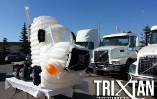 Balloon sculpture of a white semi truck displayed outdoors in front of real transport trucks at an event
